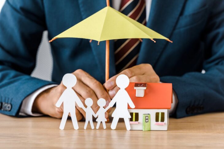 Male hands with paper cut family, house model and umbrella on wooden table
