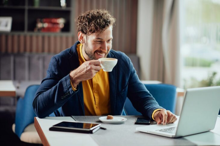 Close up of smiling freelancer sitting in cafe, drinking fresh coffee and using laptop